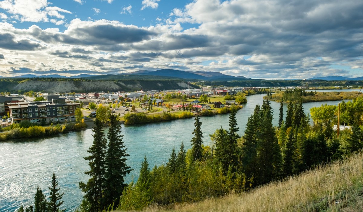 View over Whitehorse, Yukon, with the Yukon River curving past downtown beneath forested hills and a cloud-filled blue sky.